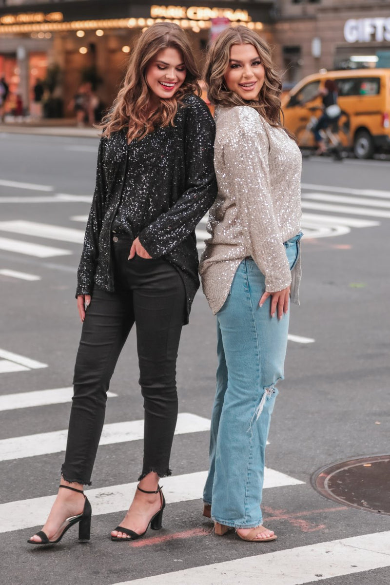 a loose long-sleeve top made of lightweight material covered in shimmering sequins with a collared neckline, fitted button closure cuffs, an accent chest pocket, and a relaxed button-up silhouette that falls into a round high-low hemline in black and champagne