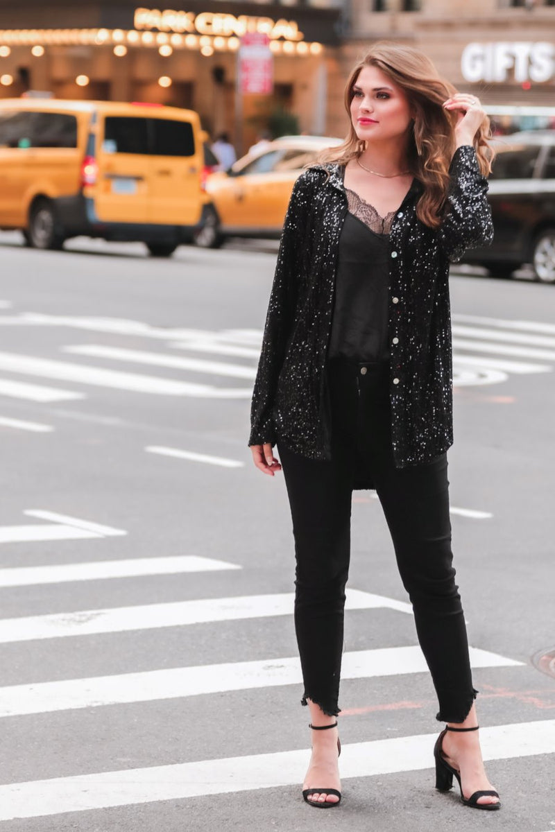 a loose long-sleeve top made of lightweight material covered in shimmering sequins with a collared neckline, fitted button closure cuffs, an accent chest pocket, and a relaxed button-up silhouette that falls into a round high-low hemline in black
