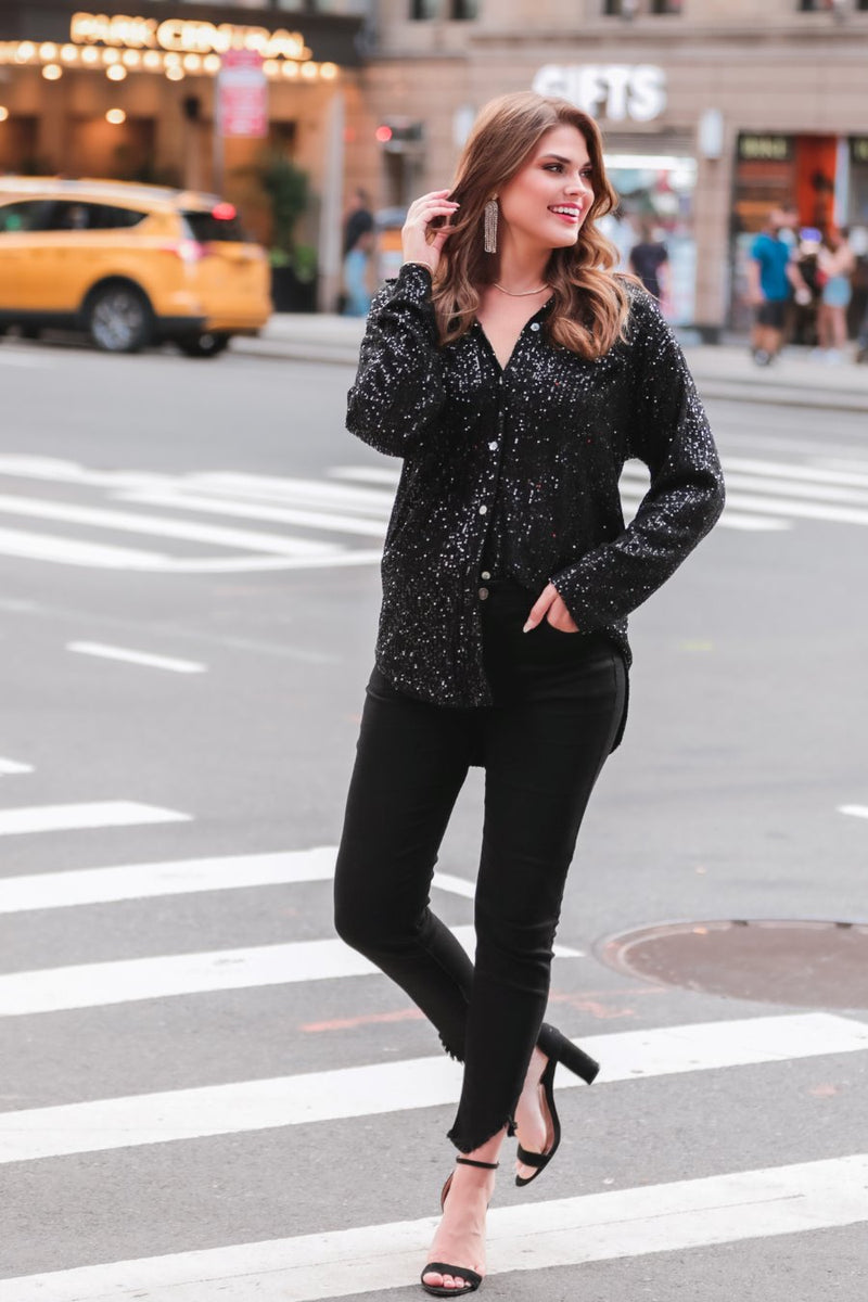 a loose long-sleeve top made of lightweight material covered in shimmering sequins with a collared neckline, fitted button closure cuffs, an accent chest pocket, and a relaxed button-up silhouette that falls into a round high-low hemline in black