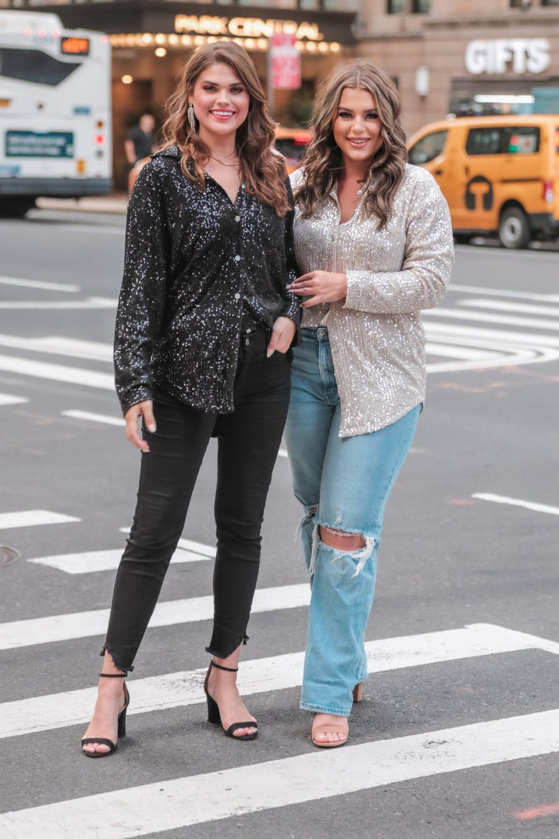 a loose long-sleeve top made of lightweight material covered in shimmering sequins with a collared neckline, fitted button closure cuffs, an accent chest pocket, and a relaxed button-up silhouette that falls into a round high-low hemline in black and champagne