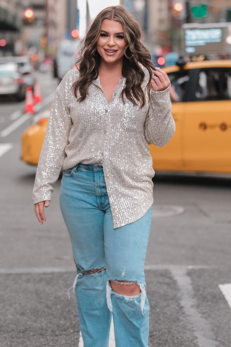 a loose long-sleeve top made of lightweight material covered in shimmering sequins with a collared neckline, fitted button closure cuffs, an accent chest pocket, and a relaxed button-up silhouette that falls into a round high-low hemline in champagne