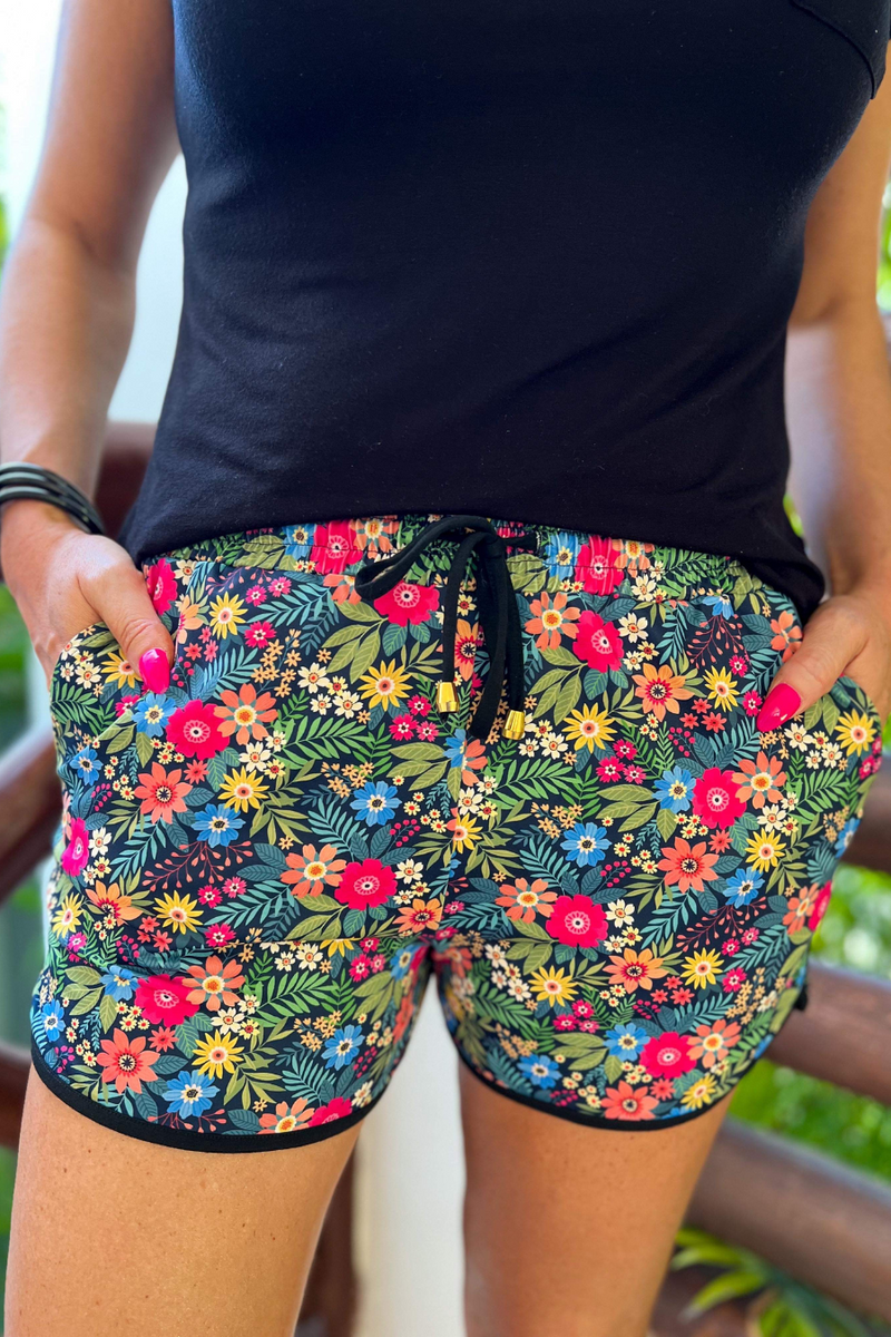 mid-rise relaxed fit activewear shorts with side pockets and a tie waist drawstring with gold bead accents, designed on a mix of multicolor floral and palm leaves pattern print fabric, navy base, and black trim