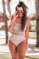 two-piece swimsuit sold separately made of stretchy material with a ruched crop top, adjustable tie straps, and removable padding paired with high-waisted bottoms on a neutral leopard print fabric