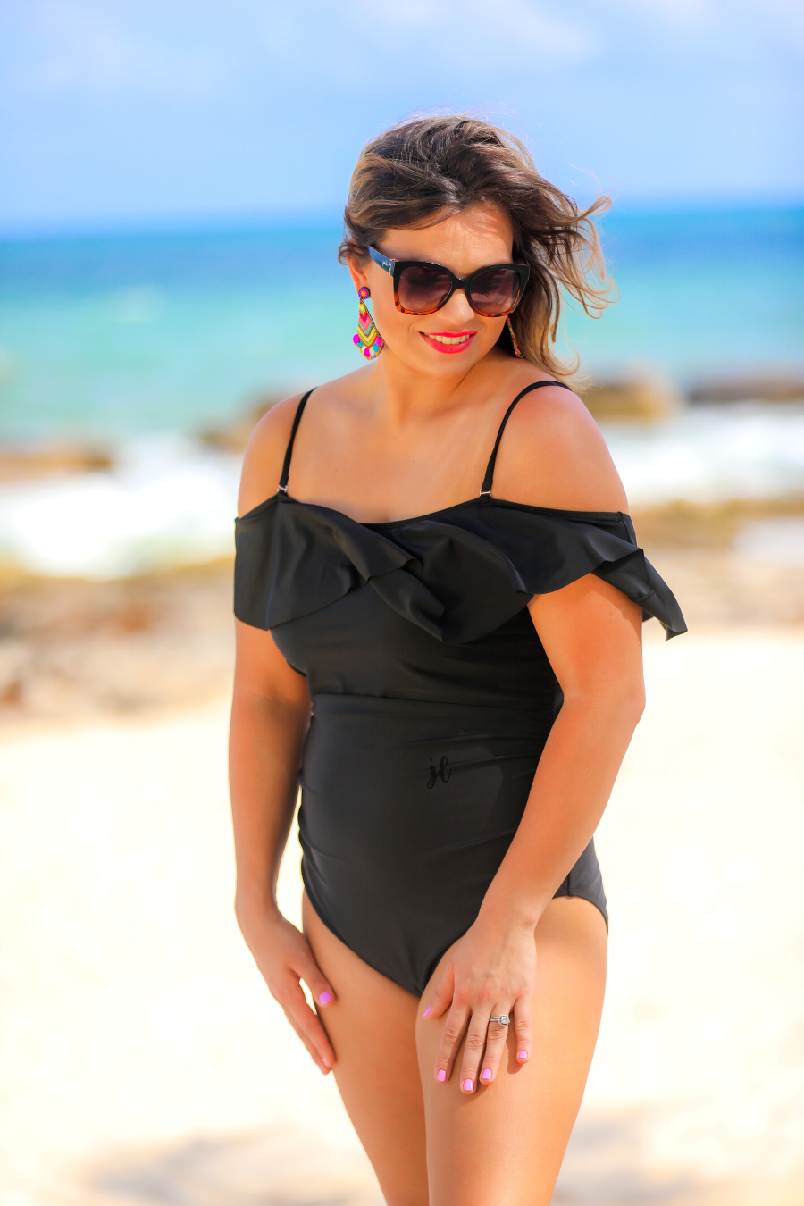 solid black one-piece swimsuit made of stretchy material, a ruffled neckline and straps that can be worn on or off the shoulder, adjustable and removable spaghetti straps, an adjustable crisscrossed cutout back, side ruched detailing, and removable padding