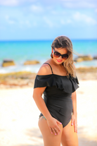 solid black one-piece swimsuit made of stretchy material, a ruffled neckline and straps that can be worn on or off the shoulder, adjustable and removable spaghetti straps, an adjustable crisscrossed cutout back, side ruched detailing, and removable padding