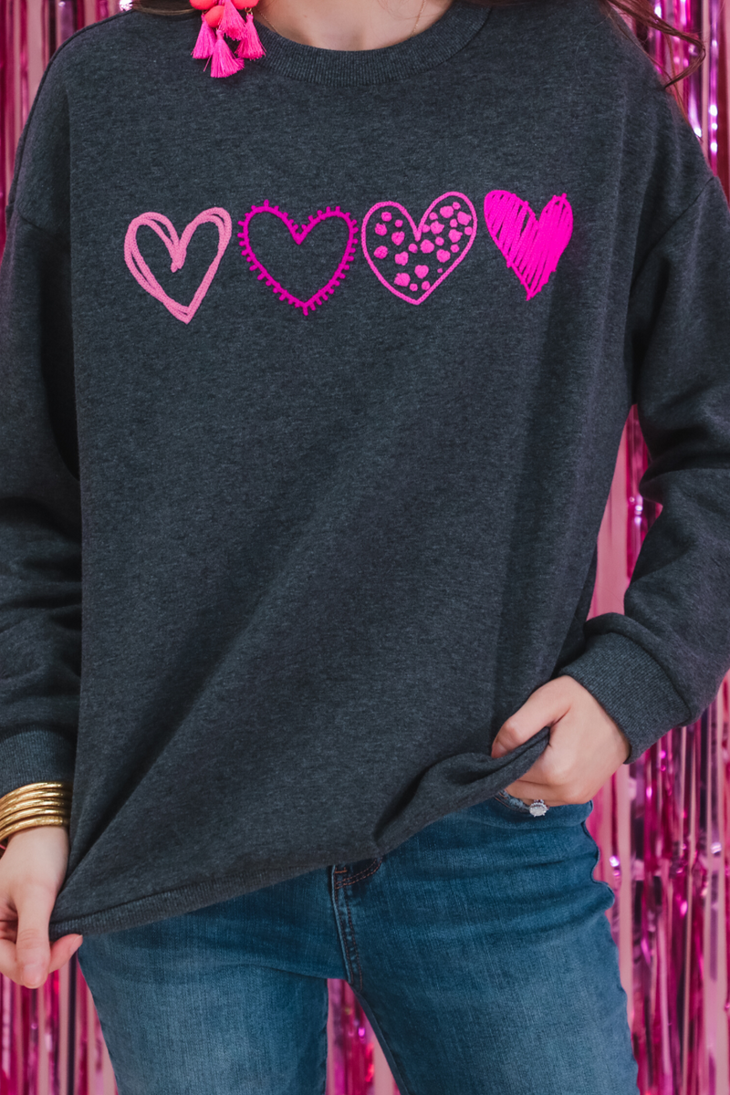 a charcoal long-sleeve sweatshirt with a fuzzy inside, sewn-on pink chenille heart patches, a relaxed drop-shoulder fit, and ribbed detailing on the classic crew neckline, cuffs, and hemline