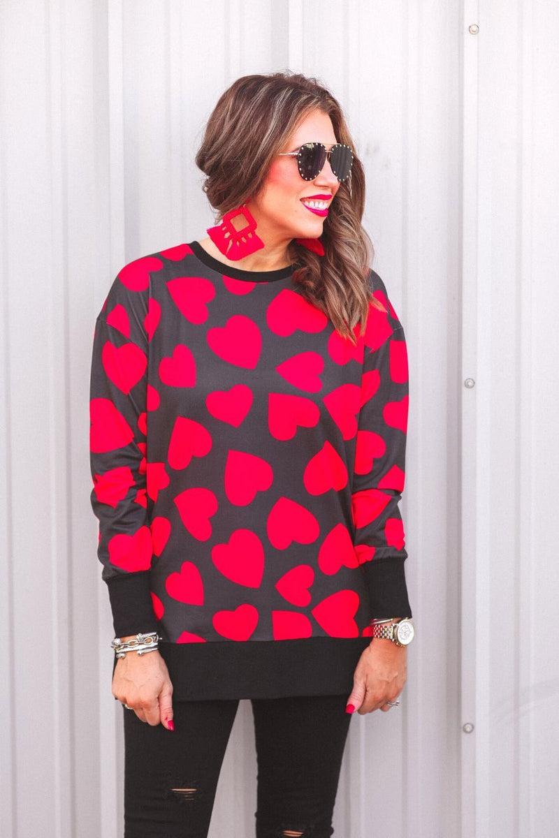 a black long-sleeve tunic with a bold red heart pattern on a black base, side slits, a relaxed drop-shoulder fit, and ribbed detailing on the neckline, cuffs, and hemline
