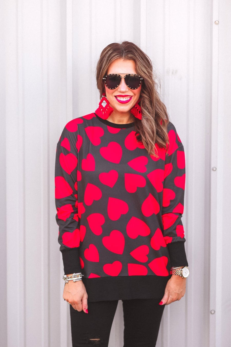 a black long-sleeve tunic with a bold red heart pattern on a black base, side slits, a relaxed drop-shoulder fit, and ribbed detailing on the neckline, cuffs, and hemline