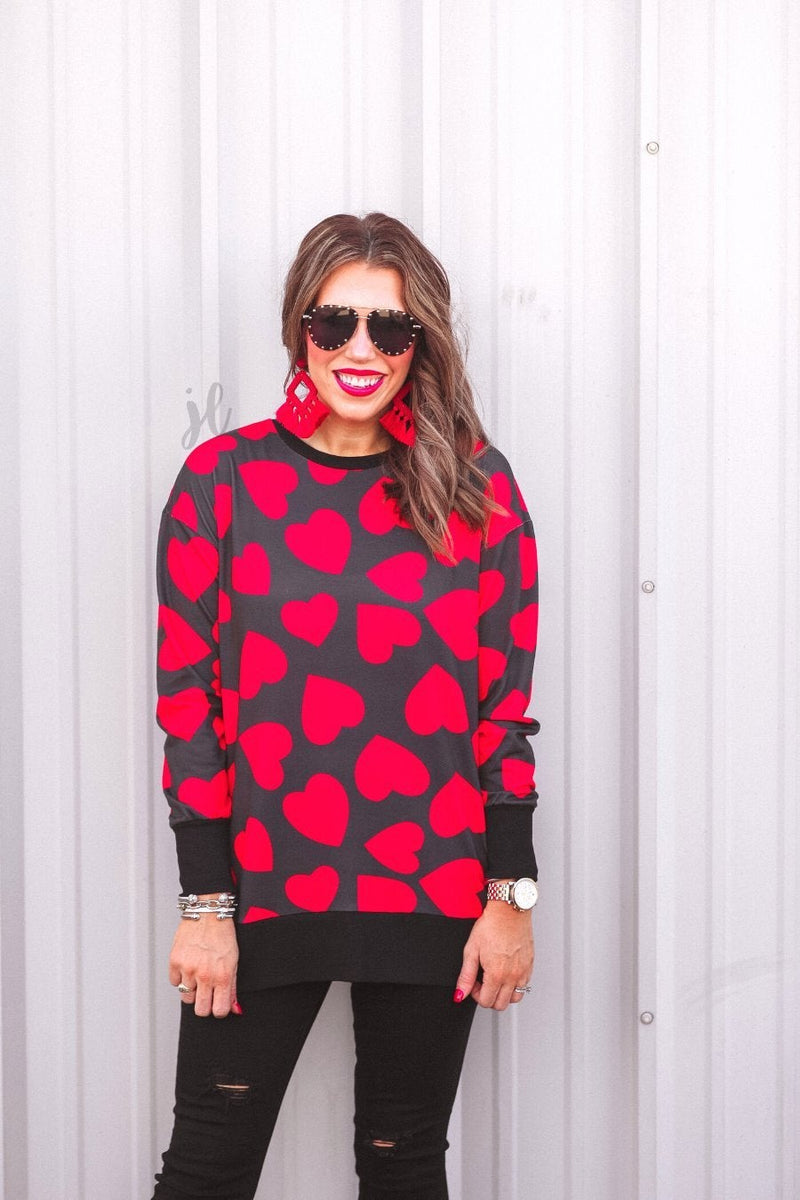 a black long-sleeve tunic with a bold red heart pattern on a black base, side slits, a relaxed drop-shoulder fit, and ribbed detailing on the neckline, cuffs, and hemline