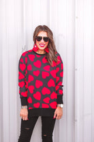 a black long-sleeve tunic with a bold red heart pattern on a black base, side slits, a relaxed drop-shoulder fit, and ribbed detailing on the neckline, cuffs, and hemline