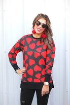a black long-sleeve tunic with a bold red heart pattern on a black base, side slits, a relaxed drop-shoulder fit, and ribbed detailing on the neckline, cuffs, and hemline