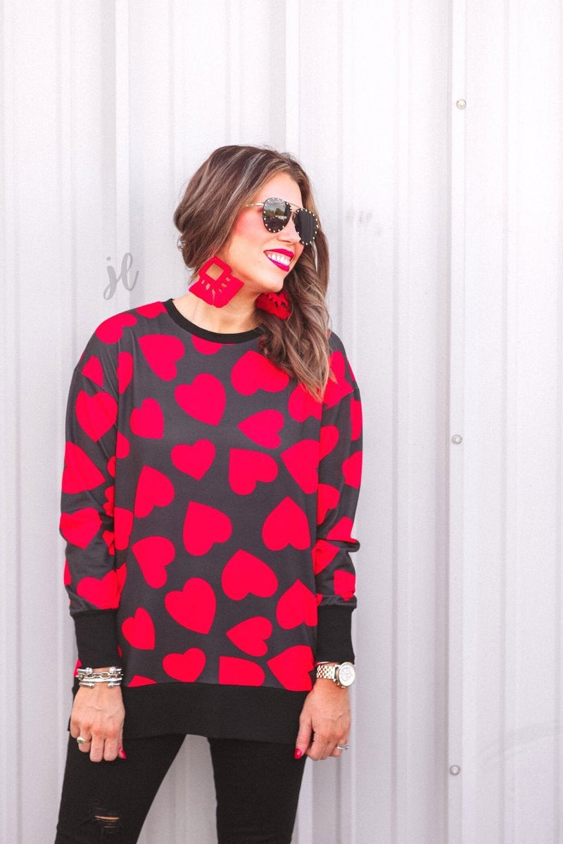 a black long-sleeve tunic with a bold red heart pattern on a black base, side slits, a relaxed drop-shoulder fit, and ribbed detailing on the neckline, cuffs, and hemline