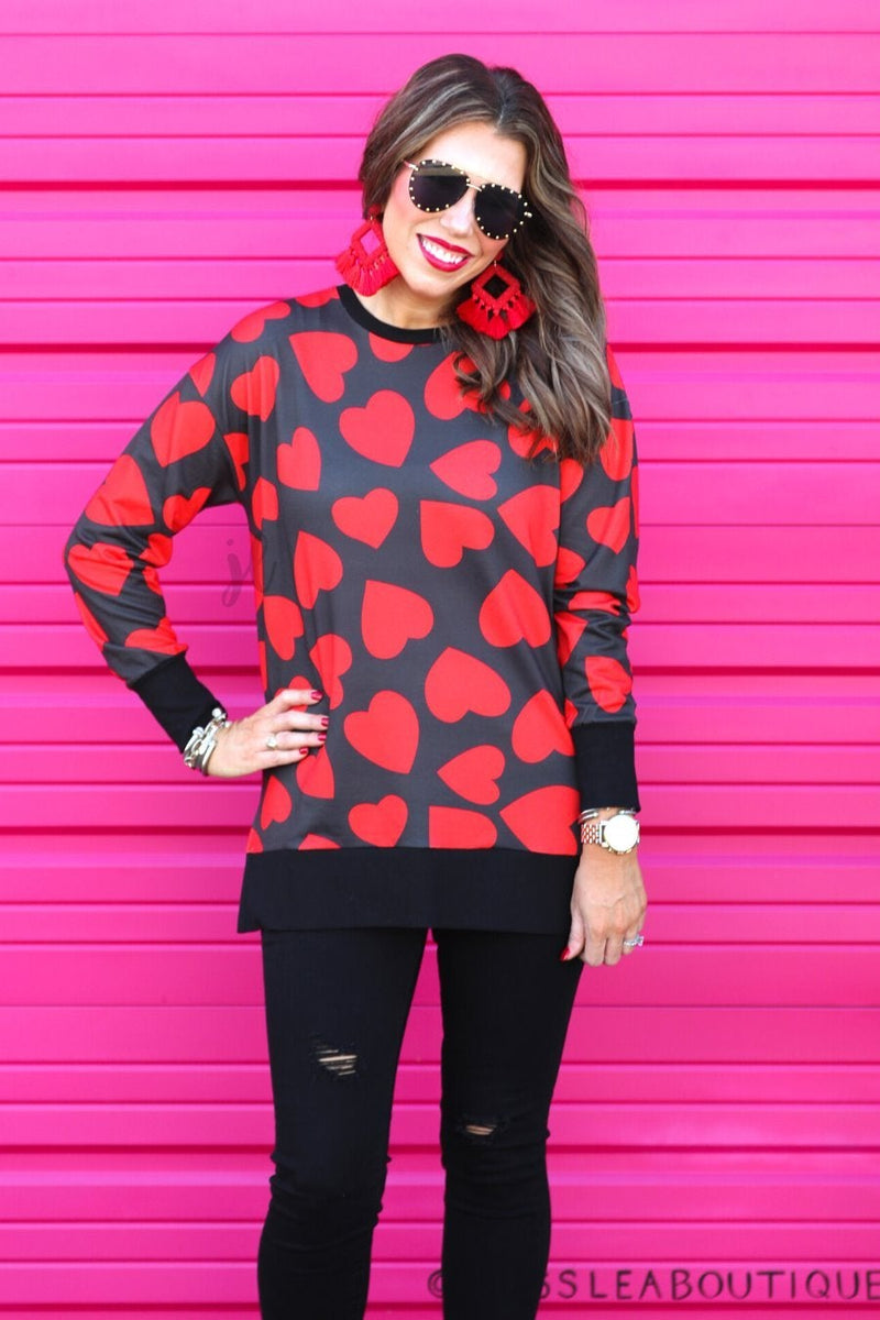 a black long-sleeve tunic with a bold red heart pattern on a black base, side slits, a relaxed drop-shoulder fit, and ribbed detailing on the neckline, cuffs, and hemline