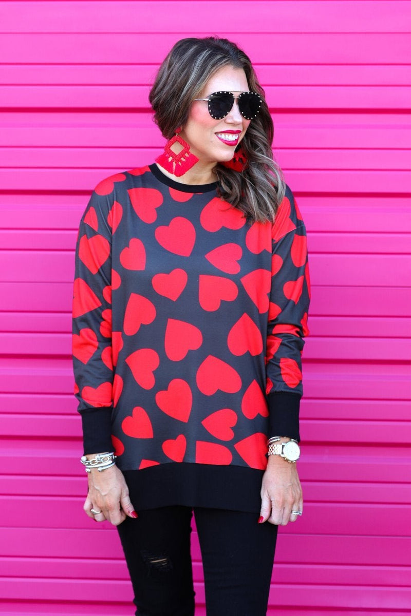 a black long-sleeve tunic with a bold red heart pattern on a black base, side slits, a relaxed drop-shoulder fit, and ribbed detailing on the neckline, cuffs, and hemline