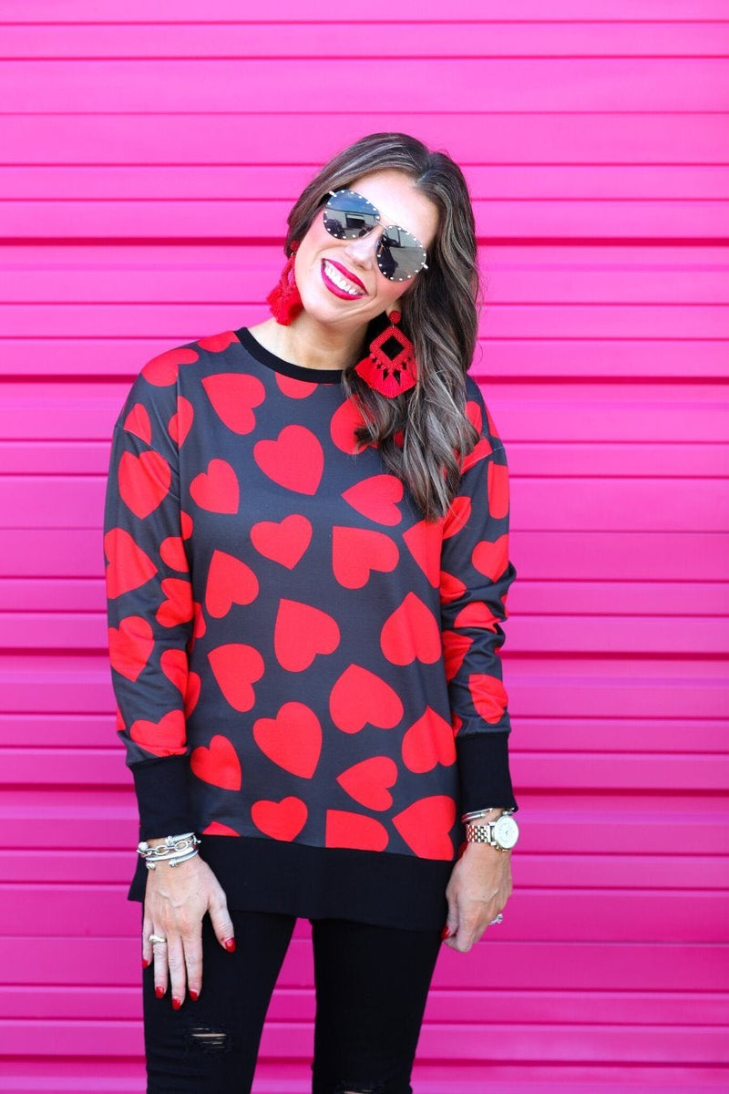 a black long-sleeve tunic with a bold red heart pattern on a black base, side slits, a relaxed drop-shoulder fit, and ribbed detailing on the neckline, cuffs, and hemline