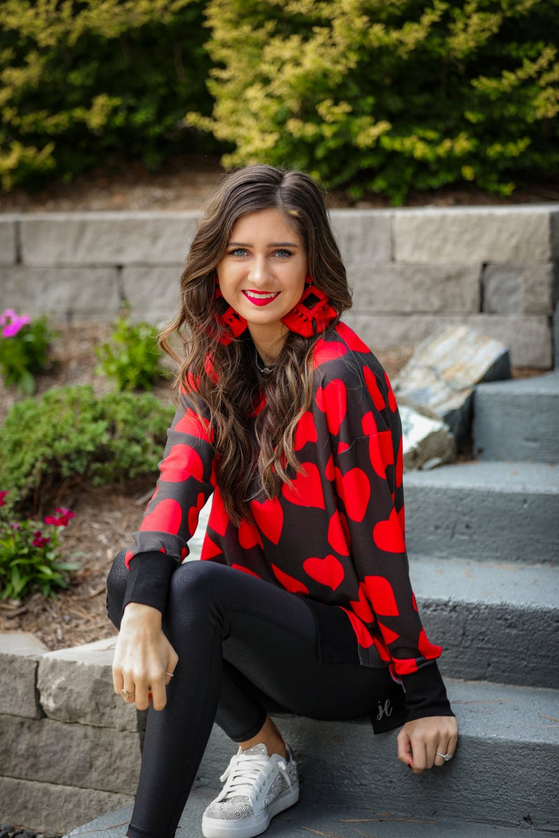 a black long-sleeve tunic with a bold red heart pattern on a black base, side slits, a relaxed drop-shoulder fit, and ribbed detailing on the neckline, cuffs, and hemline