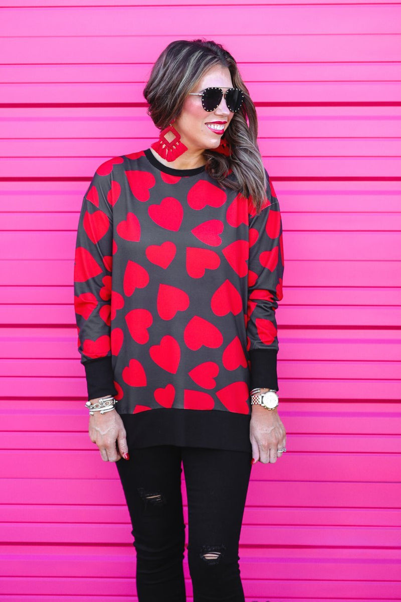 a black long-sleeve tunic with a bold red heart pattern on a black base, side slits, a relaxed drop-shoulder fit, and ribbed detailing on the neckline, cuffs, and hemline