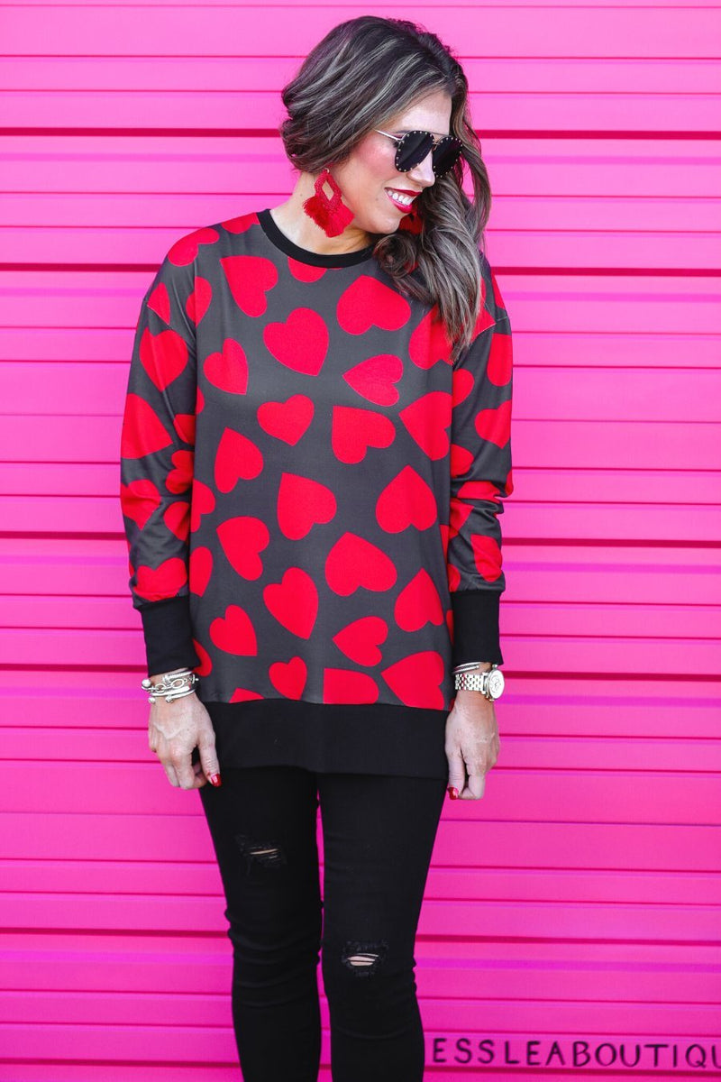 a black long-sleeve tunic with a bold red heart pattern on a black base, side slits, a relaxed drop-shoulder fit, and ribbed detailing on the neckline, cuffs, and hemline