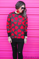 a black long-sleeve tunic with a bold red heart pattern on a black base, side slits, a relaxed drop-shoulder fit, and ribbed detailing on the neckline, cuffs, and hemline