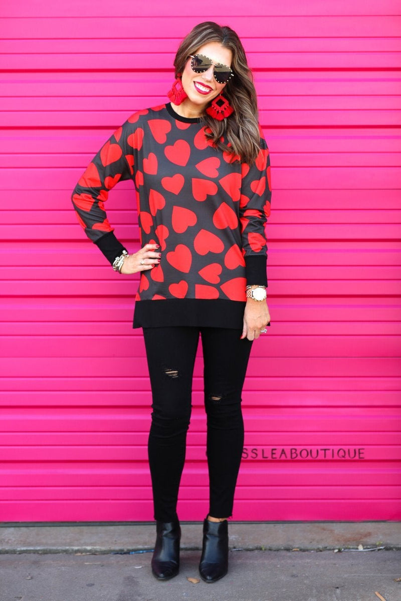 a black long-sleeve tunic with a bold red heart pattern on a black base, side slits, a relaxed drop-shoulder fit, and ribbed detailing on the neckline, cuffs, and hemline