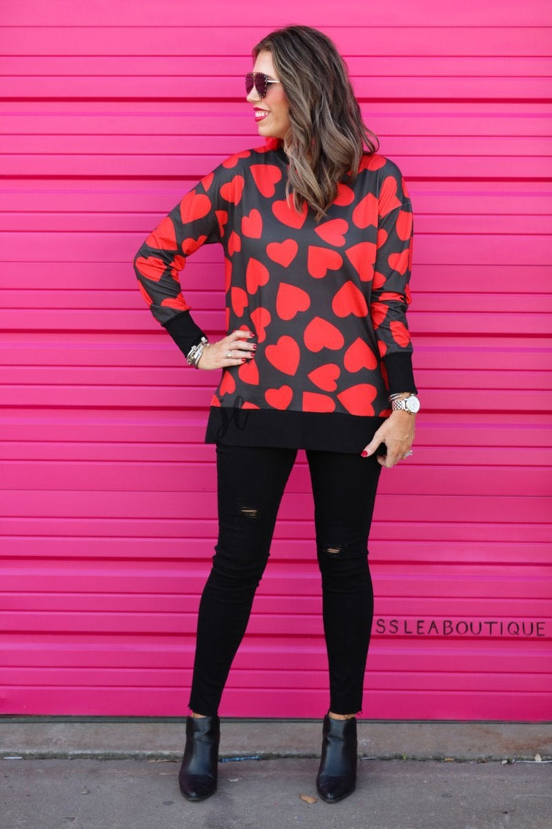 a black long-sleeve tunic with a bold red heart pattern on a black base, side slits, a relaxed drop-shoulder fit, and ribbed detailing on the neckline, cuffs, and hemline