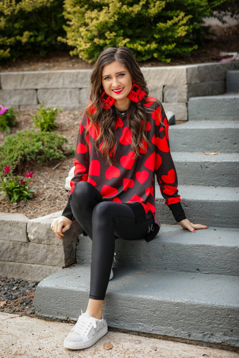 a black long-sleeve tunic with a bold red heart pattern on a black base, side slits, a relaxed drop-shoulder fit, and ribbed detailing on the neckline, cuffs, and hemline