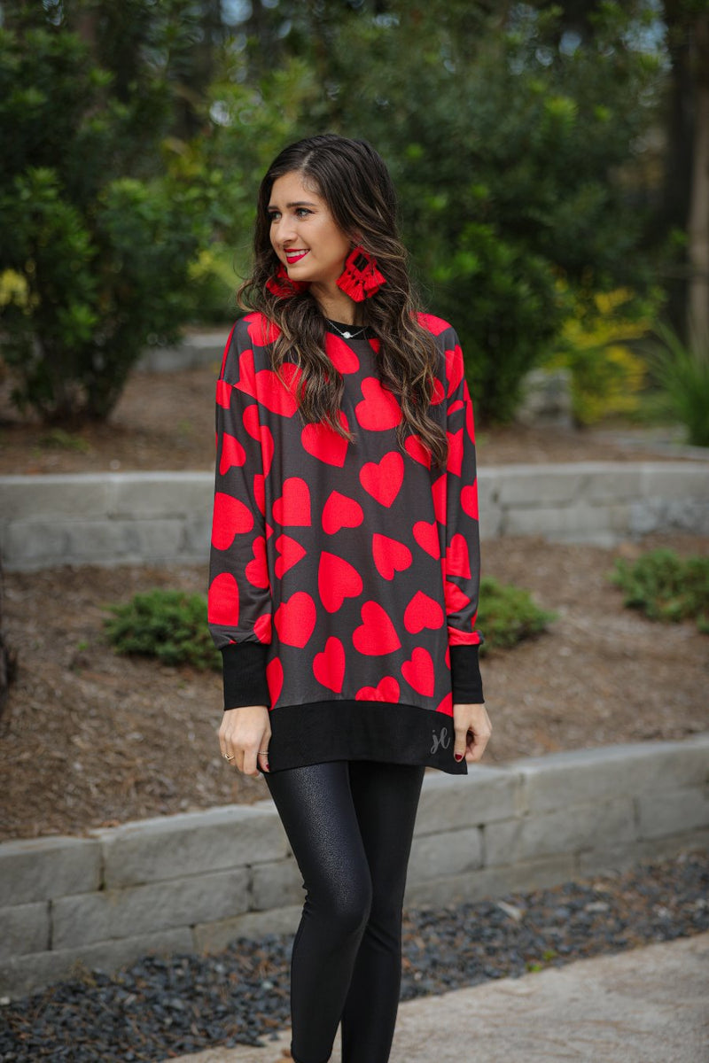 a black long-sleeve tunic with a bold red heart pattern on a black base, side slits, a relaxed drop-shoulder fit, and ribbed detailing on the neckline, cuffs, and hemline