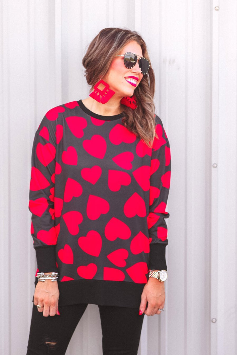 a black long-sleeve tunic with a bold red heart pattern on a black base, side slits, a relaxed drop-shoulder fit, and ribbed detailing on the neckline, cuffs, and hemline