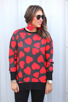 a black long-sleeve tunic with a bold red heart pattern on a black base, side slits, a relaxed drop-shoulder fit, and ribbed detailing on the neckline, cuffs, and hemline