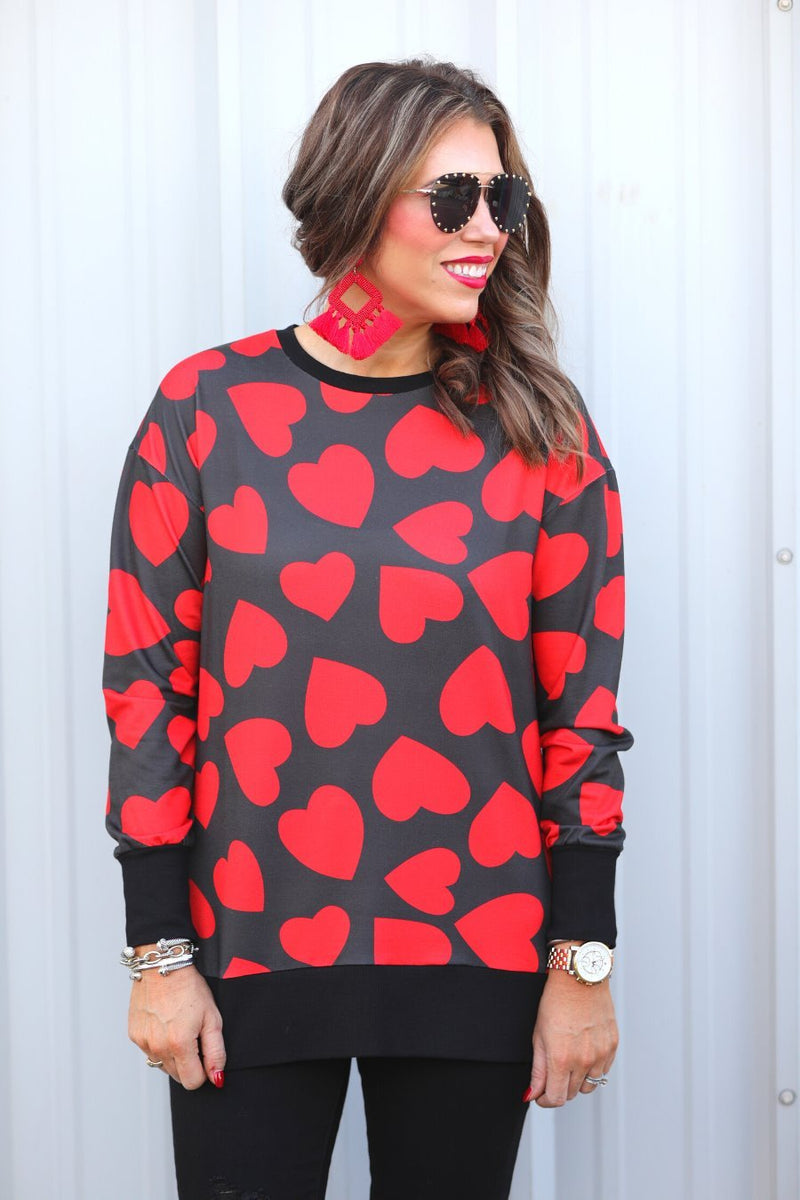 a black long-sleeve tunic with a bold red heart pattern on a black base, side slits, a relaxed drop-shoulder fit, and ribbed detailing on the neckline, cuffs, and hemline