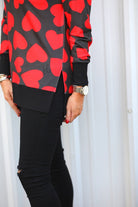 a black long-sleeve tunic with a bold red heart pattern on a black base, side slits, a relaxed drop-shoulder fit, and ribbed detailing on the neckline, cuffs, and hemline