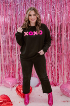 a black long-sleeve sweatshirt with a soft fleece interior, a drop-shoulder silhouette, ribbed detailing on the classic crew neckline, cuffs, and hemline, finished with bold pink sewn-on chenille 'XOXO' patches