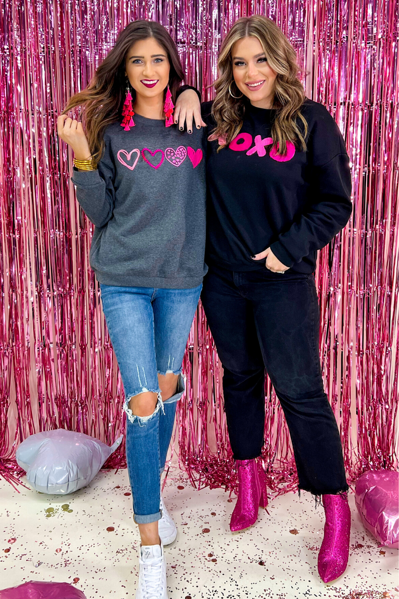 a black long-sleeve sweatshirt with a soft fleece interior, a drop-shoulder silhouette, ribbed detailing on the classic crew neckline, cuffs, and hemline, finished with bold pink sewn-on chenille 'XOXO' patches