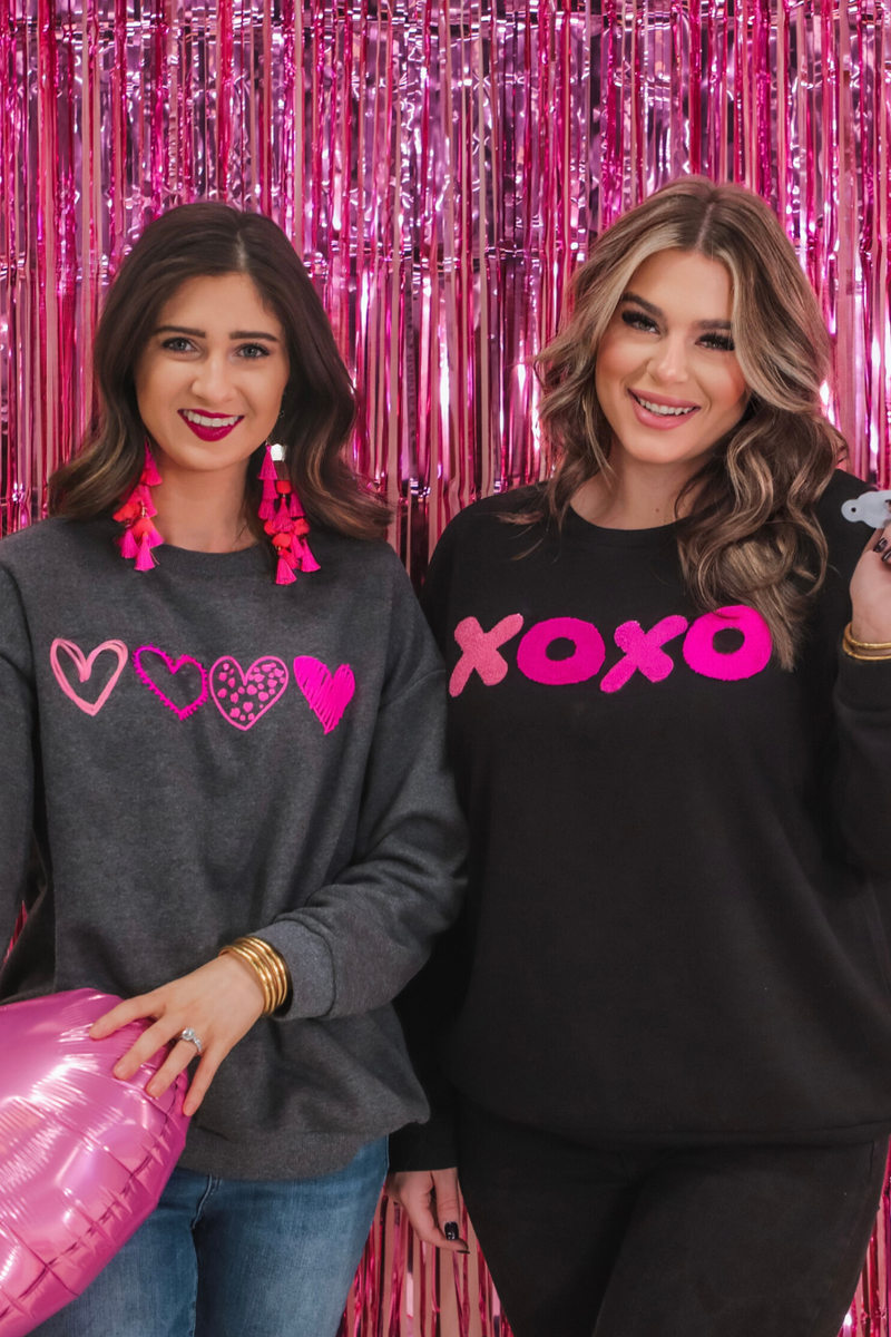 a black long-sleeve sweatshirt with a soft fleece interior, a drop-shoulder silhouette, ribbed detailing on the classic crew neckline, cuffs, and hemline, finished with bold pink sewn-on chenille 'XOXO' patches