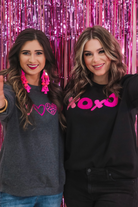 a black long-sleeve sweatshirt with a soft fleece interior, a drop-shoulder silhouette, ribbed detailing on the classic crew neckline, cuffs, and hemline, finished with bold pink sewn-on chenille 'XOXO' patches