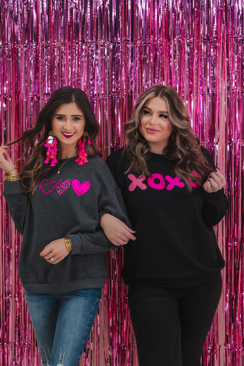 a black long-sleeve sweatshirt with a soft fleece interior, a drop-shoulder silhouette, ribbed detailing on the classic crew neckline, cuffs, and hemline, finished with bold pink sewn-on chenille 'XOXO' patches